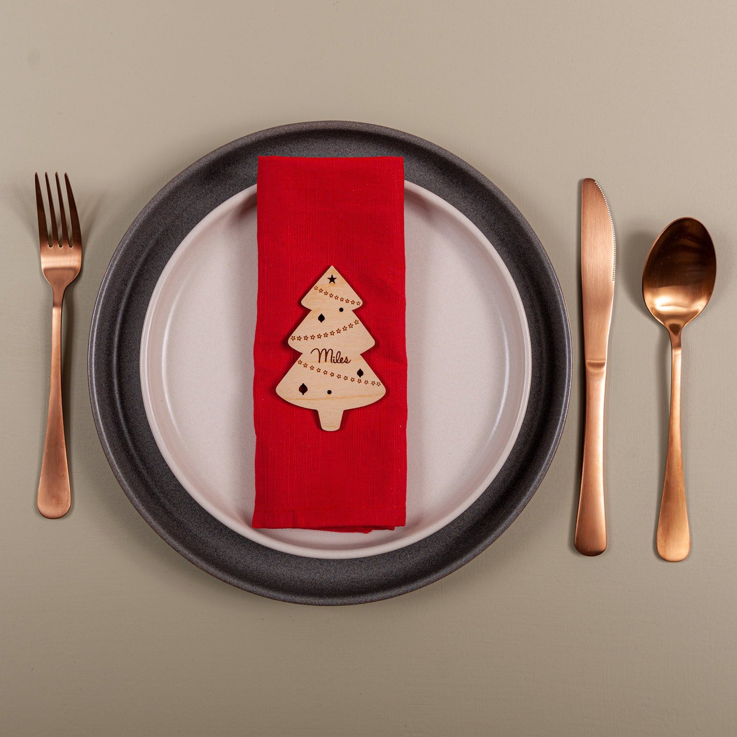 Dinner setting with a red napkin featuring a Christmas tree design, black plate, rose gold cutlery on a beige background.