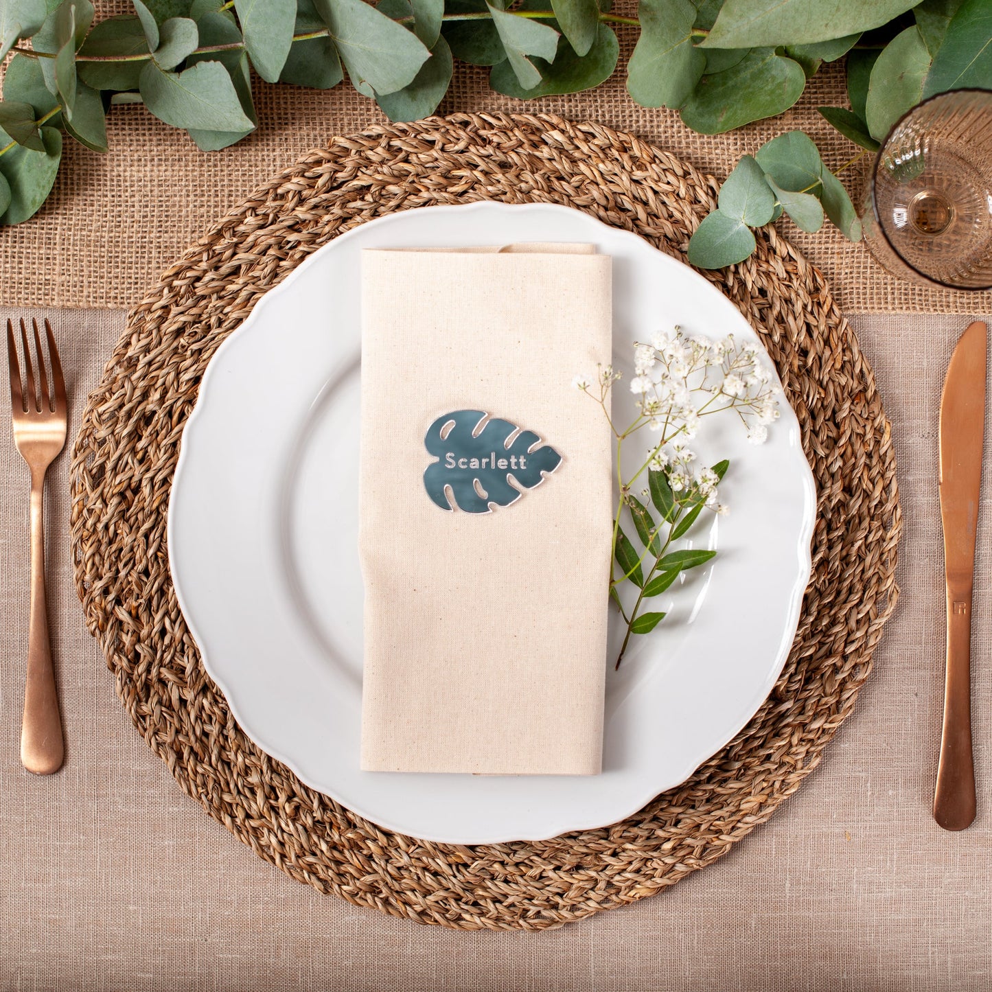 Table setting with a woven placemat, white plate, beige napkin, and cutlery on a brown tablecloth.