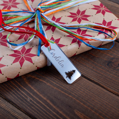 Gift wrapped in brown paper with red star patterns, colorful ribbons, and a name tag with christmas tree cut-out on a wooden surface.