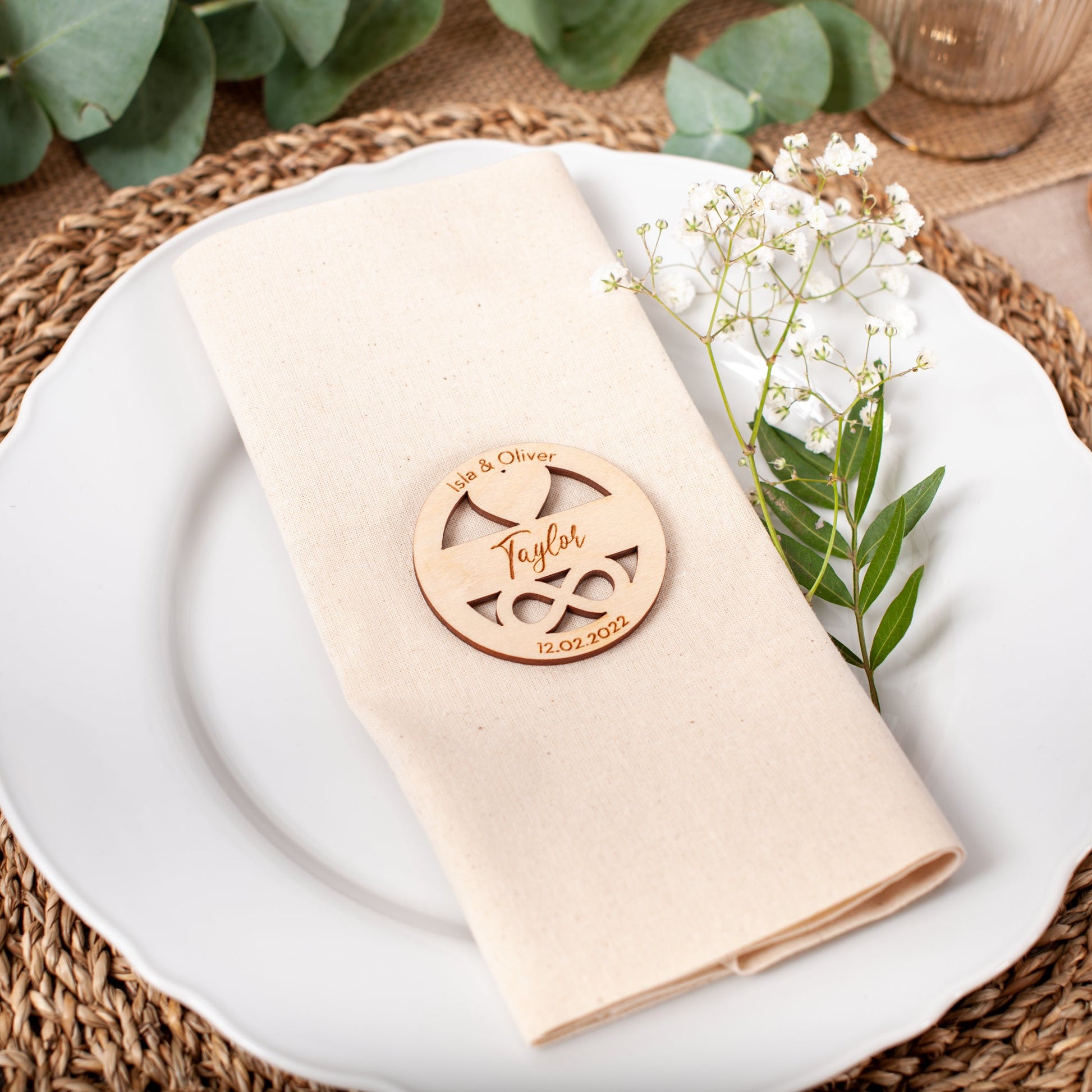 Decorative place setting with a wooden name card on a white plate with a beige napkin, surrounded by greenery.