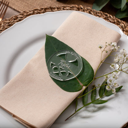 Beige napkin with a decorative leaf and emblem on a white plate with greenery, placed on a woven mat.