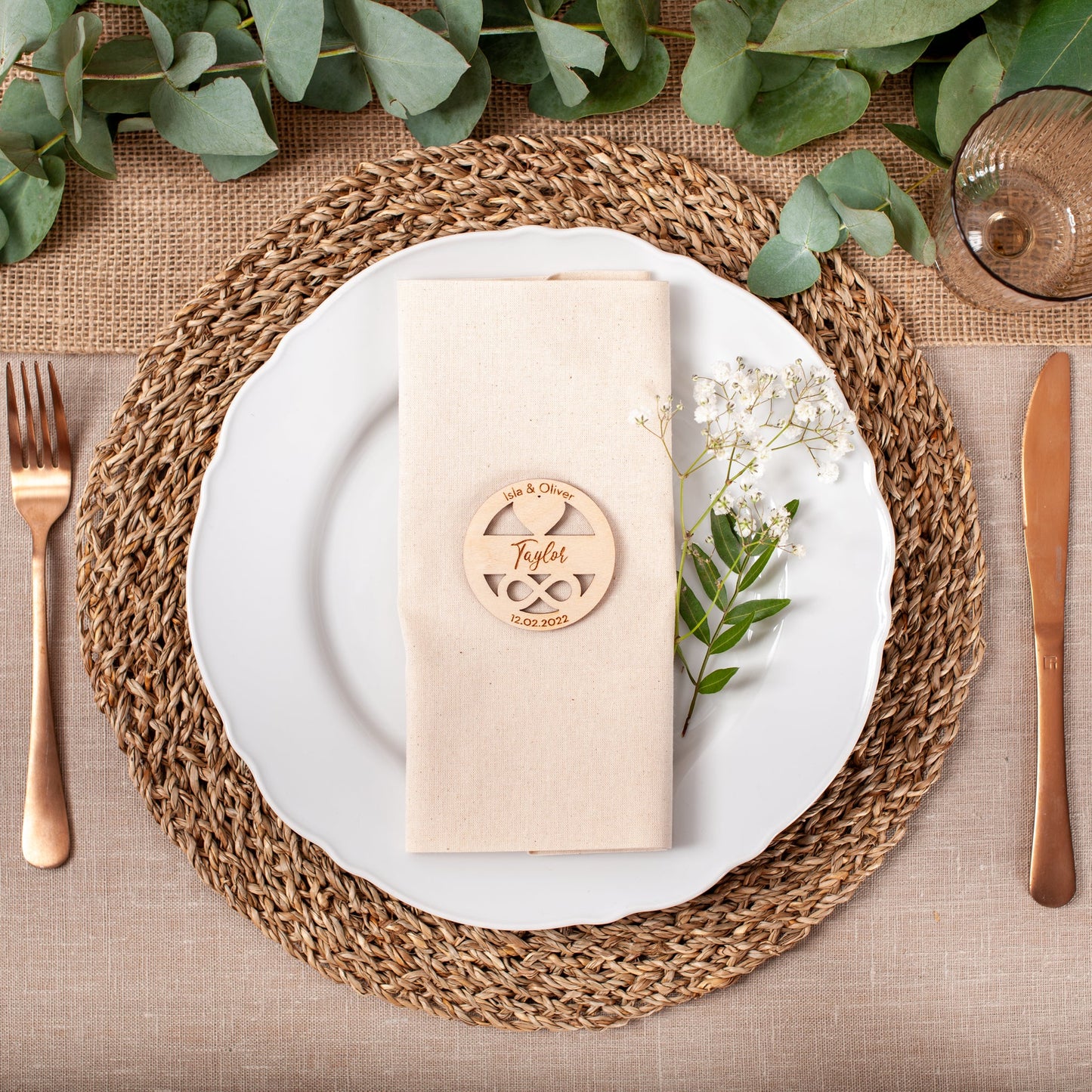 Table setting with a woven placemat, white plate, gold fork and knife, and a beige napkin with a wooden name card.
