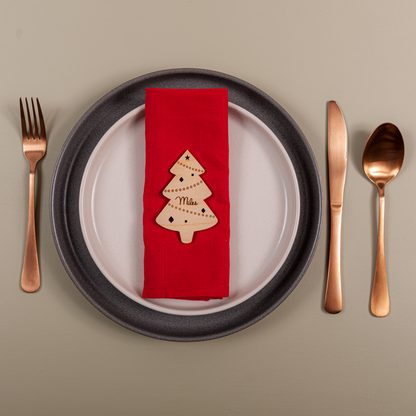 Dinner setting with a red napkin featuring a Christmas tree design, black plate, rose gold cutlery on a beige background.