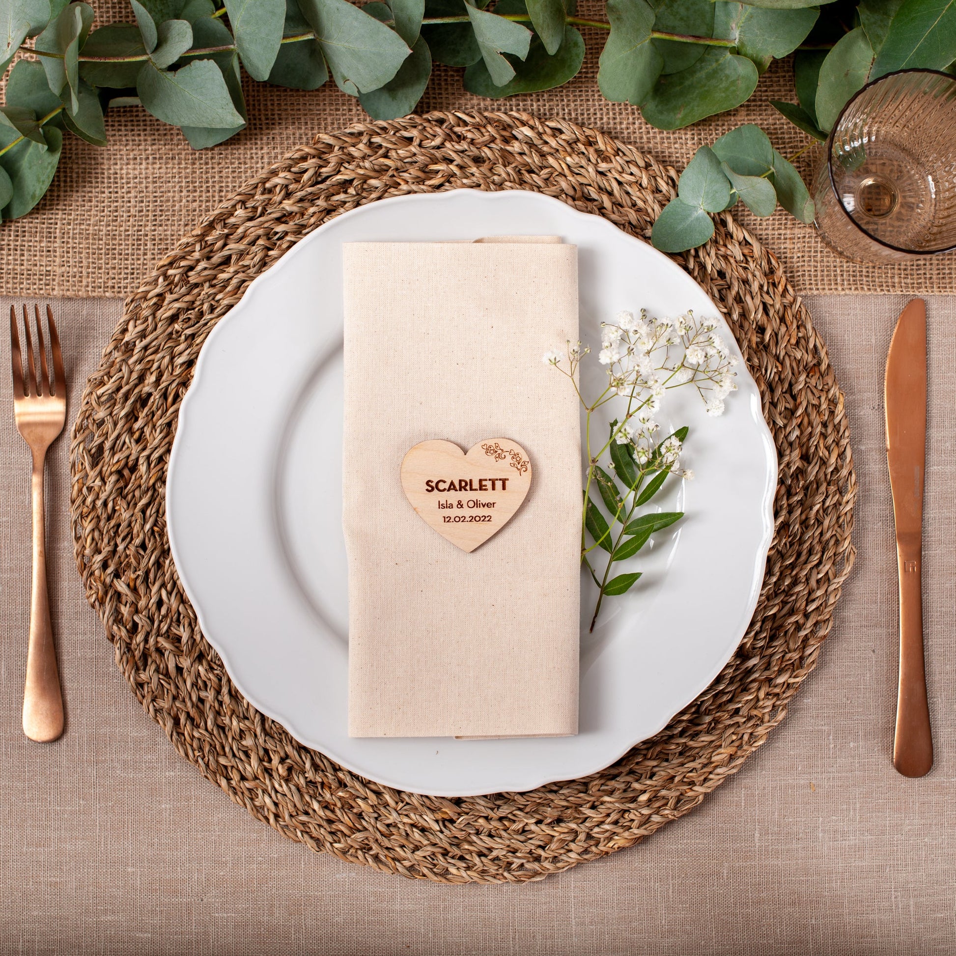 Table setting with a heart-shaped name card on a beige napkin, surrounded by cutlery and greenery.