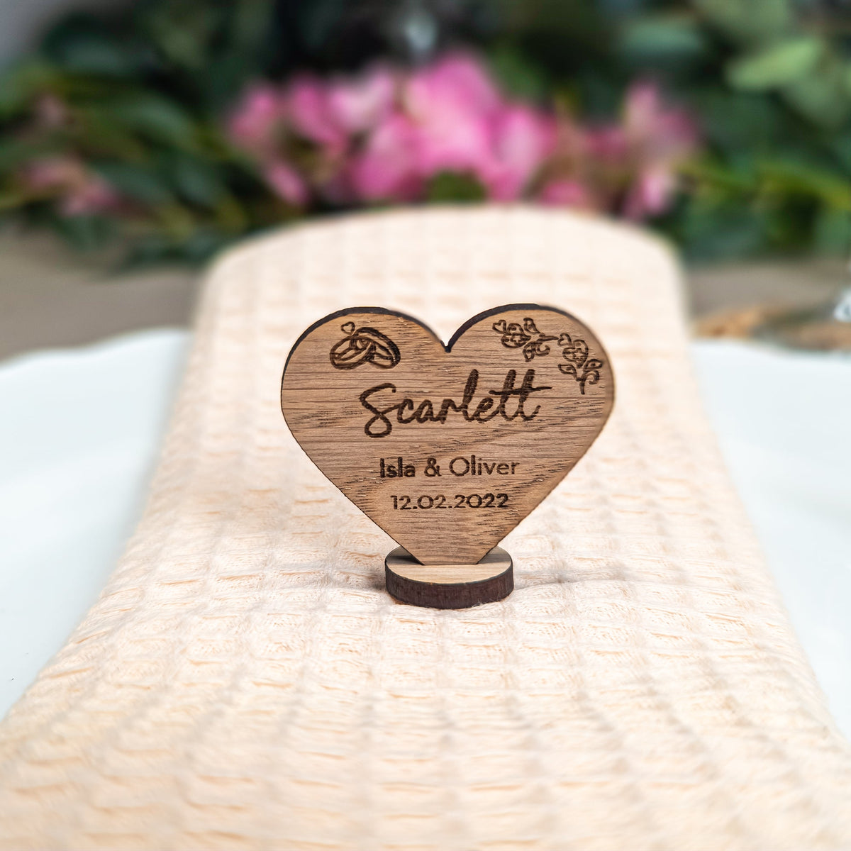 Wooden heart-shaped place card with engraved text on a woven mat with flowers in the background