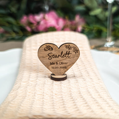 Wooden heart-shaped ornament with engraved text on a woven mat with flowers and a glass in the background.