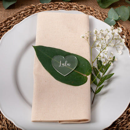 Table setting with a white plate, beige napkin, green leaf, heart-shaped card, and small white flowers on a woven placemat.