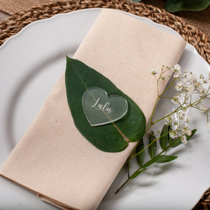 Plated setting with a folded beige napkin, green leaf, heart-shaped name card, and white flowers on a woven placemat.