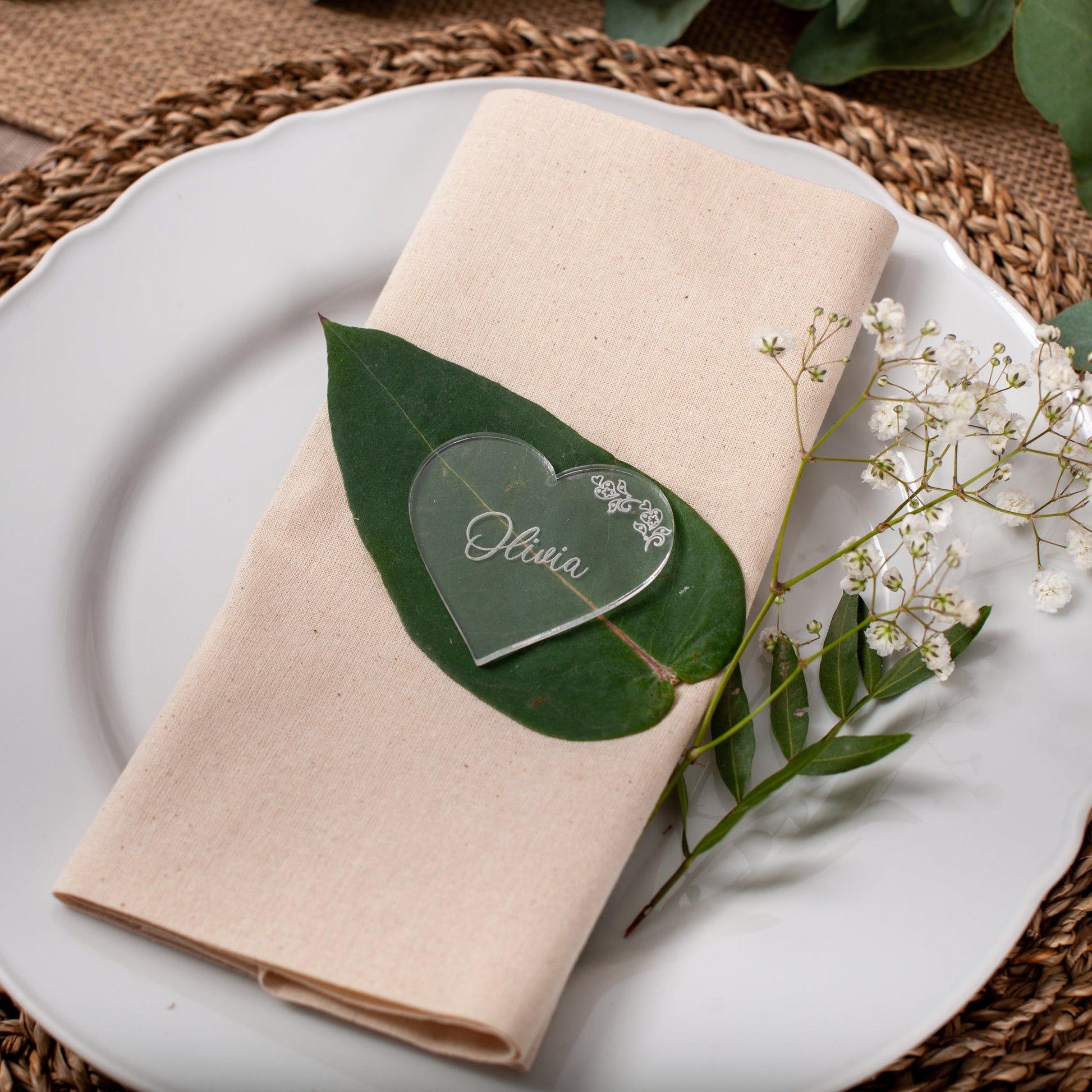 Place setting with beige napkin, green leaf card, and white flowers on a woven placemat.