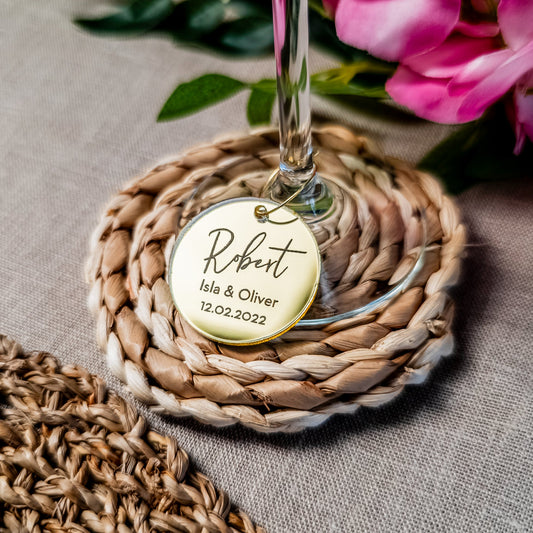 Personalized wine glass charm with a name and date on a woven coaster with flowers in the background.