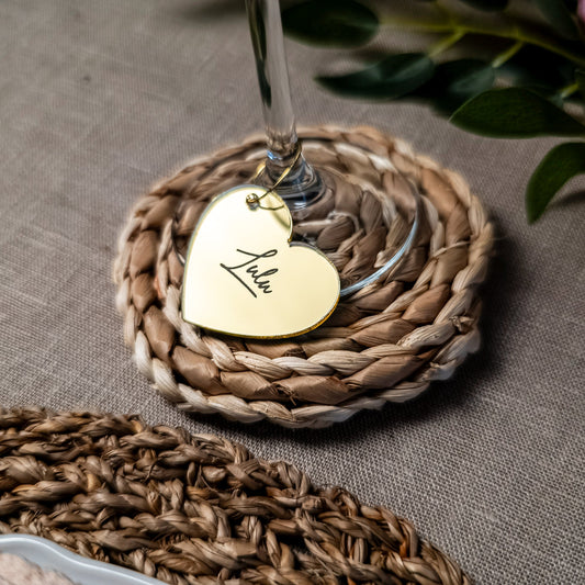 Woven coaster with a gold heart-shaped tag on a glass, placed on a textured surface.