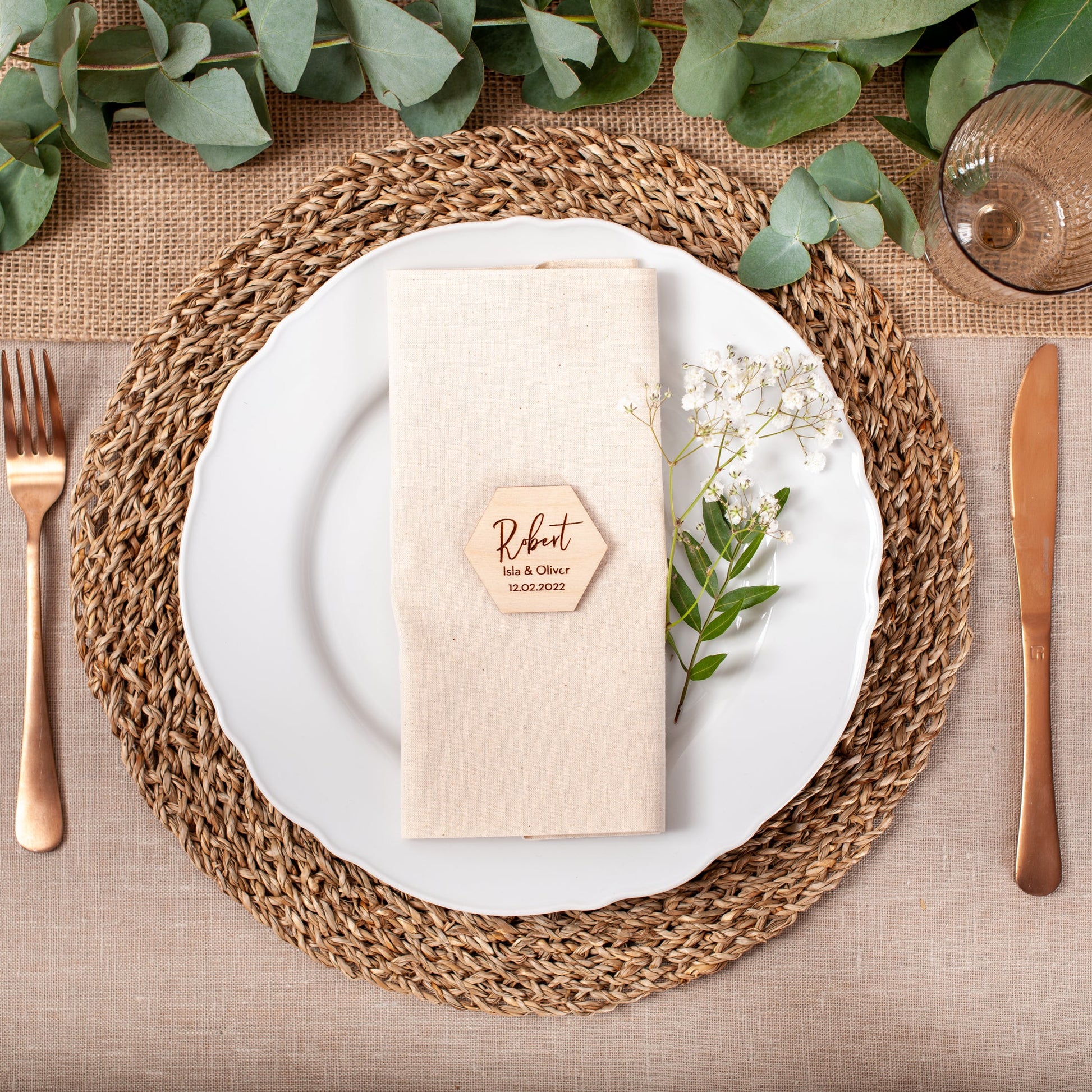Table setting with white plate, woven placemat, gold cutlery, and greenery.