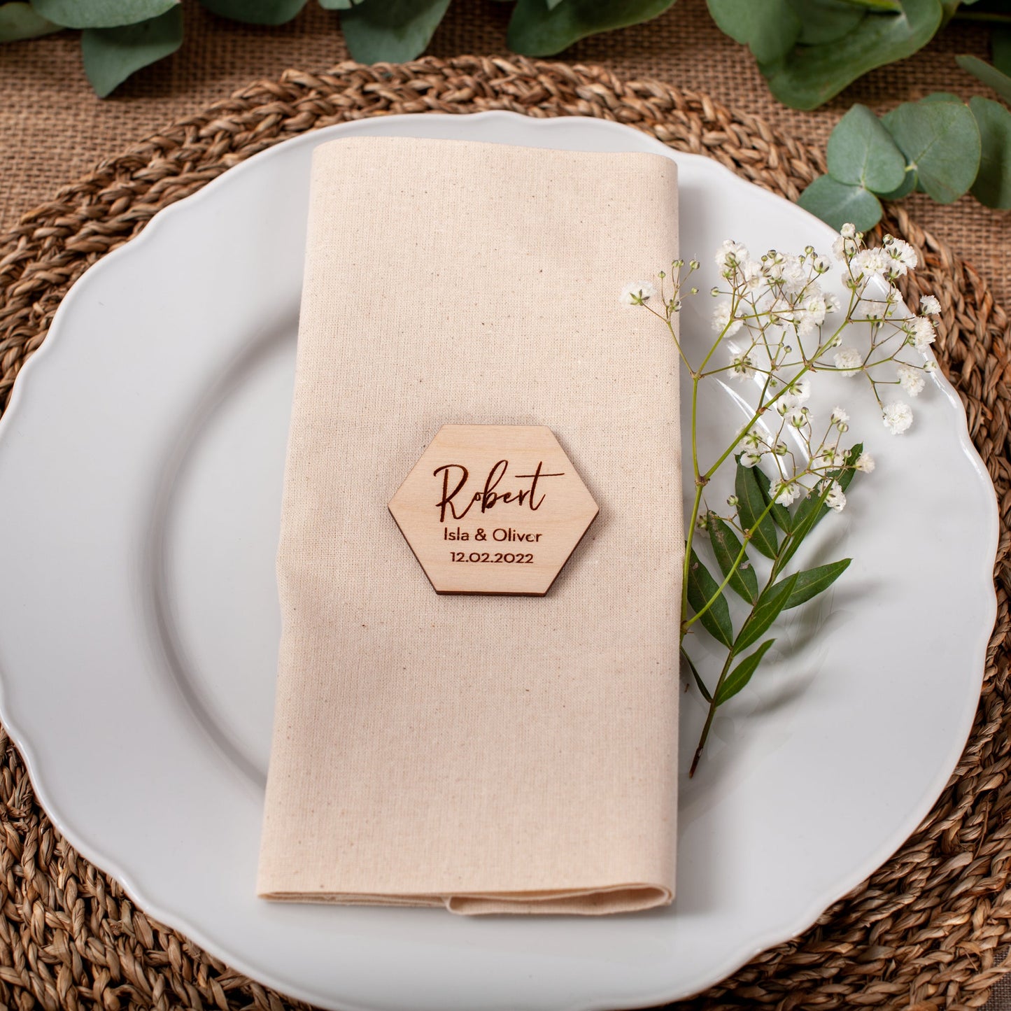 Place setting with a folded beige napkin, wooden name card, and white flowers on a woven placemat.