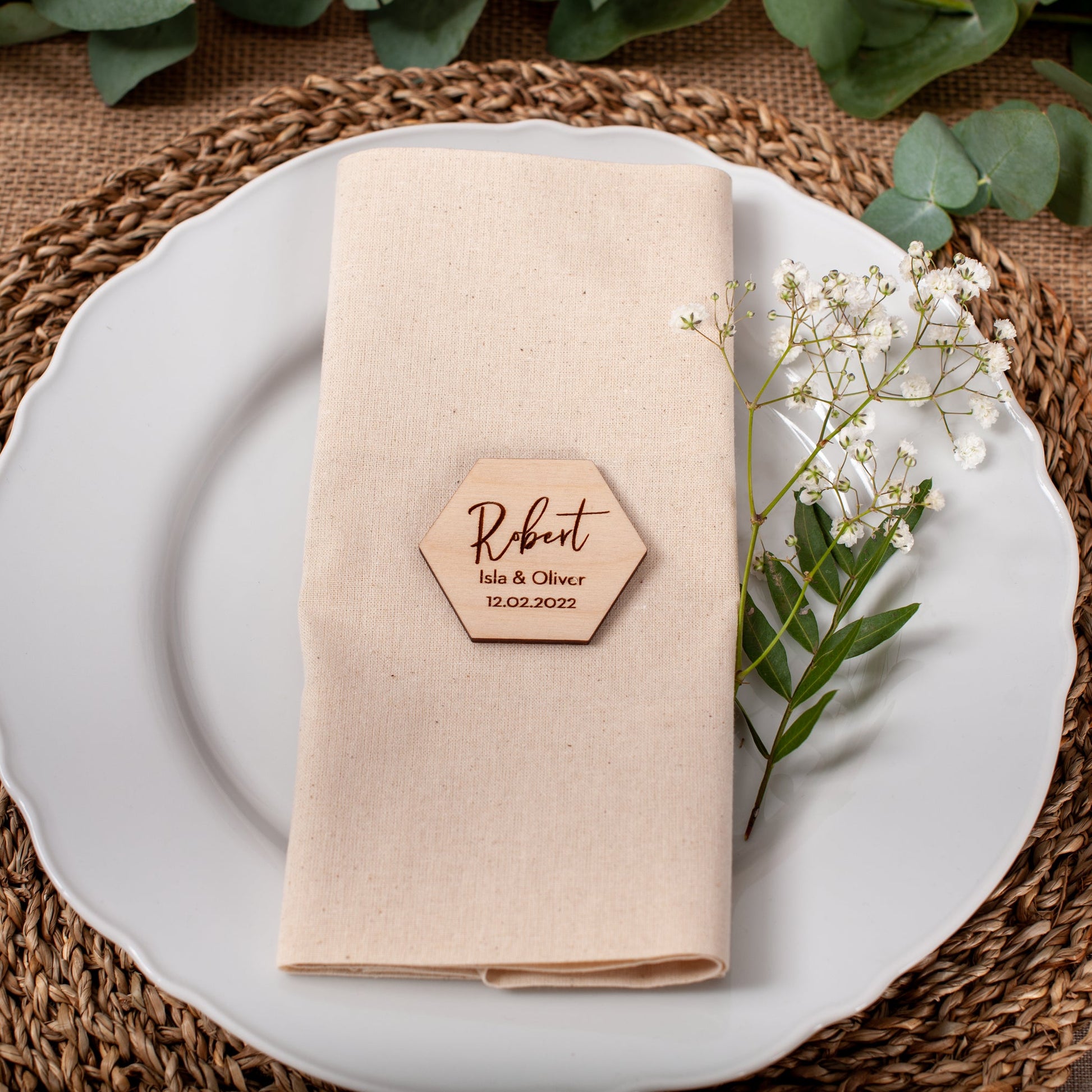 Place setting with a folded beige napkin, wooden name card, and white flowers on a woven placemat.