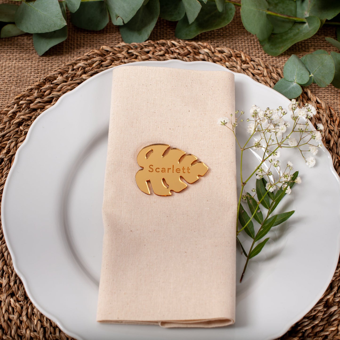 Table setting with a folded beige napkin, gold leaf name card, and white flowers on a woven placemat.