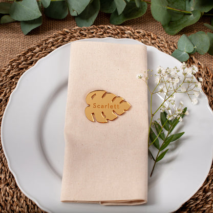 Table setting with a folded beige napkin, gold leaf name card, and white flowers on a woven placemat.