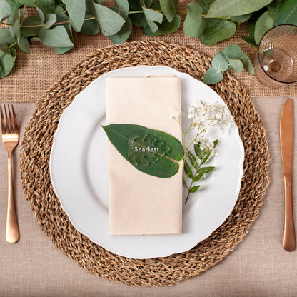 Elegant table setting with a woven placemat, white plate, gold cutlery, and greenery.